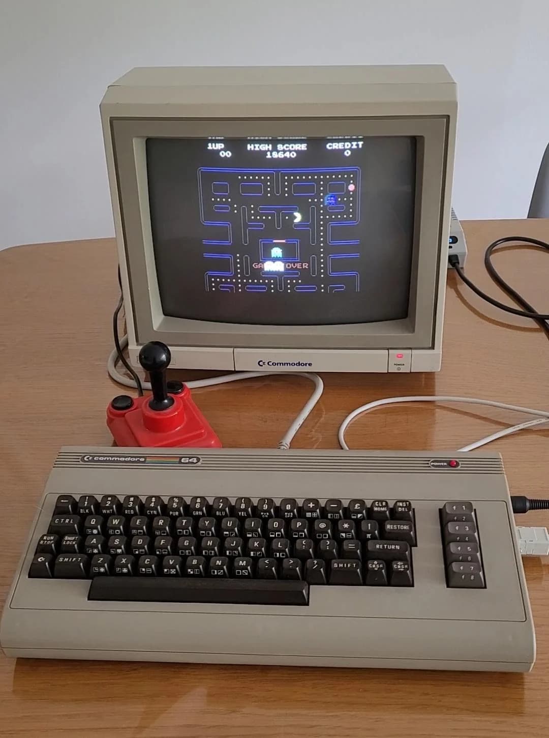 A Commodore 64 with its keyboard, a red joystick and a CRT running Pac-Man